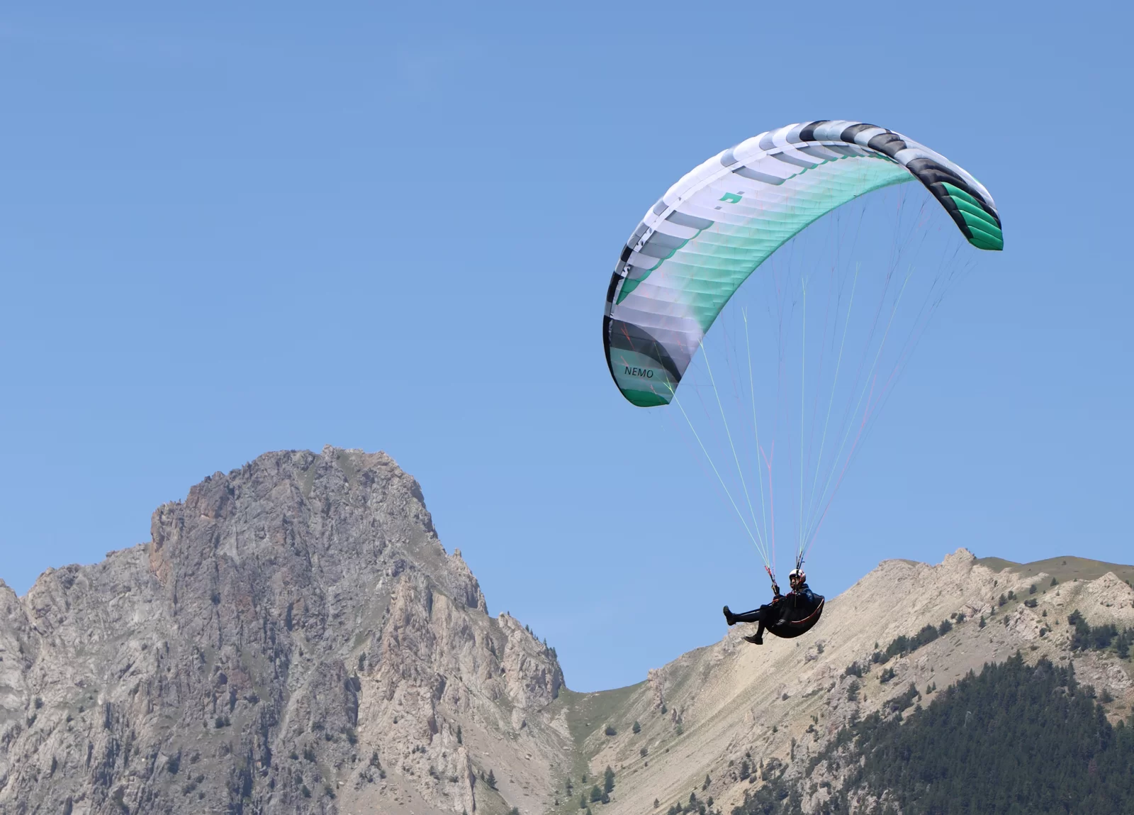 Green and white Nemo 5 paraglider flying above a rugged mountain peak under a clear blue sky