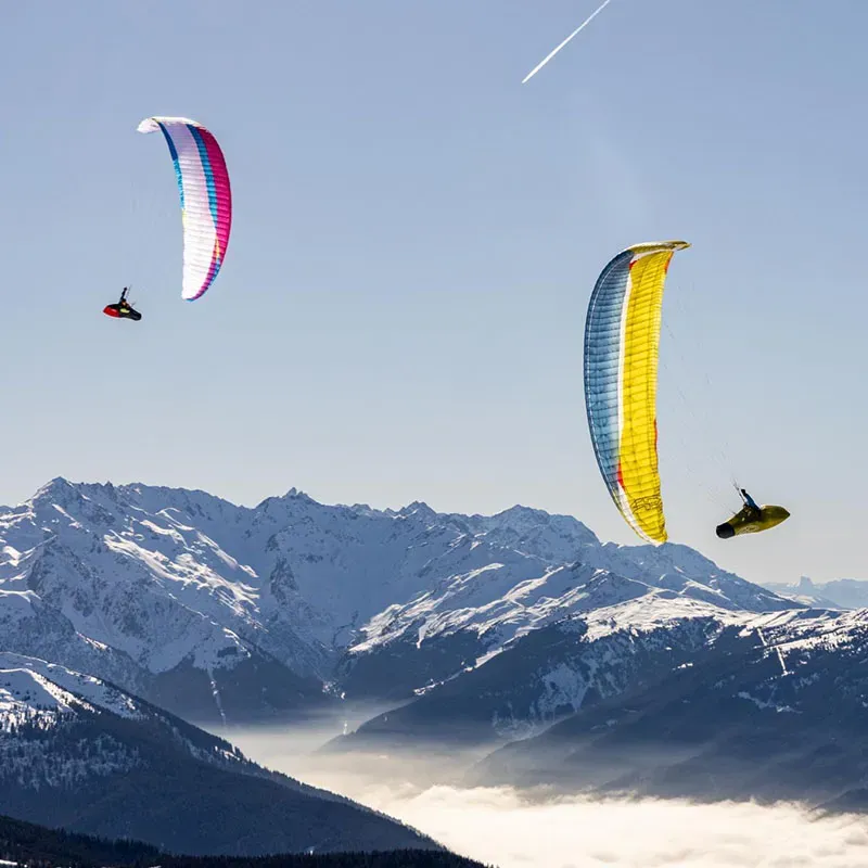 Pink and yellow paraglider canopy flying high above a snowy mountain range with a distant pilot visible