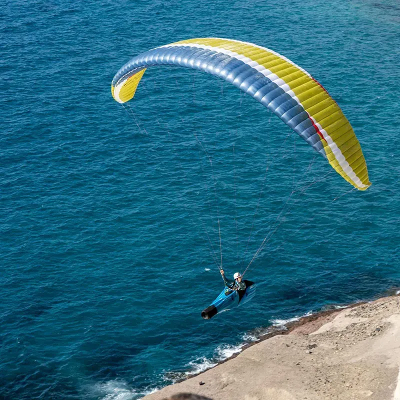 Yellow and blue paraglider wing soaring above deep blue water with a distant coastline