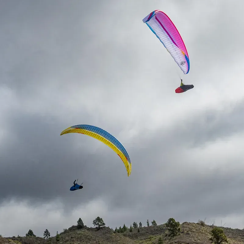 Pink and blue paraglider flying through a cloudy sky above a mountainous landscape