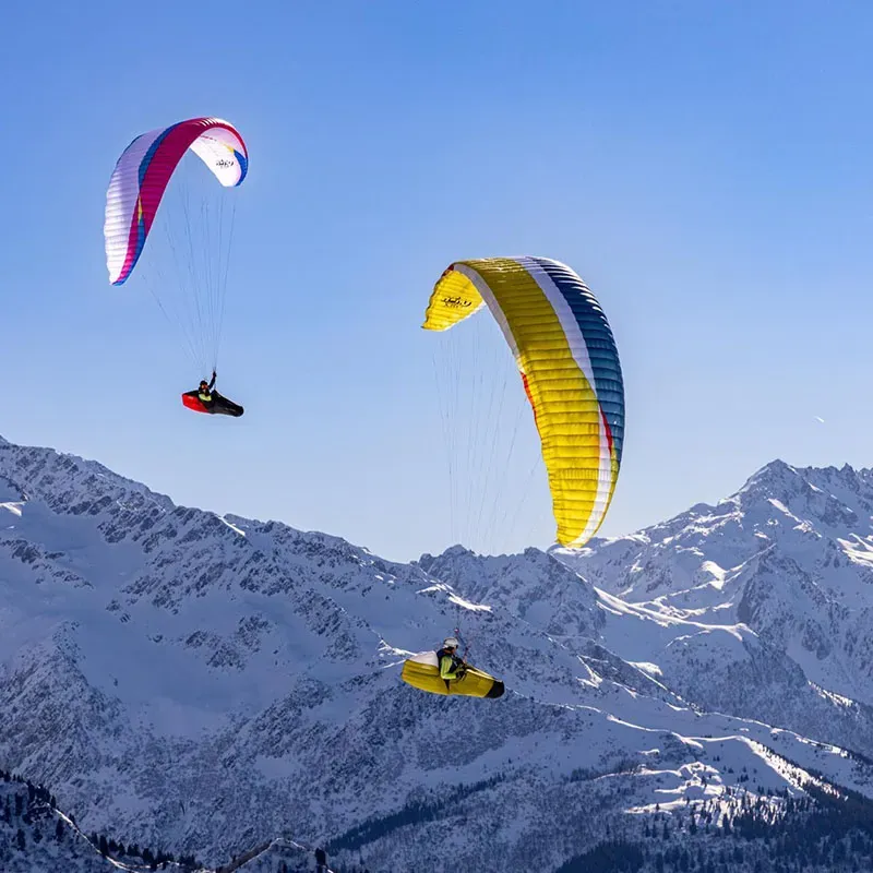 Pink and yellow Airdesign Hero Xped paraglider flying above snow-covered mountain terrain