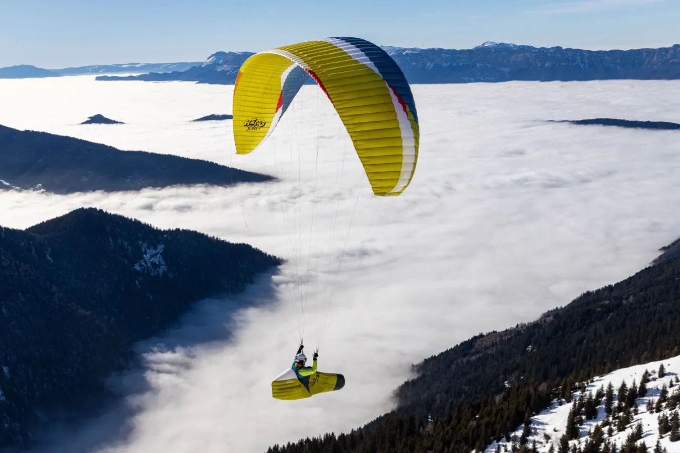 Yellow and blue paraglider wing flying above a snow-covered landscape with dark mountain peaks