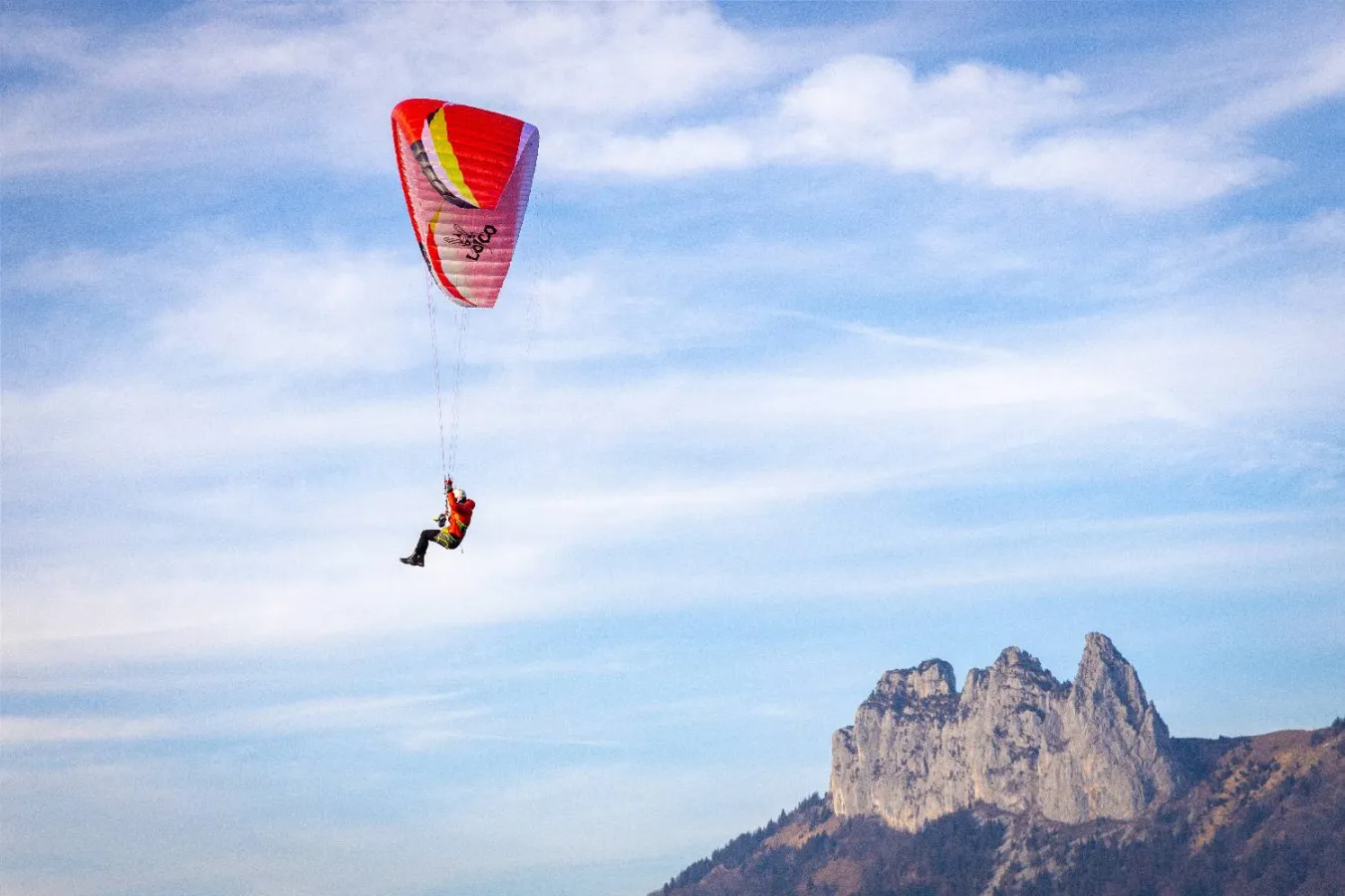 Red and yellow Airdesign Loco paraglider flying high against a blue sky with scattered clouds